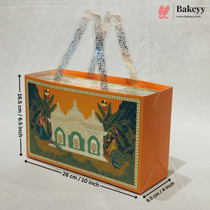 An orange rectangular gift box with clear handles and a printed traditional Indian architectural illustration on the front, shown with dimension arrows and labels indicating 26 cm length, 16.5 cm height, and 9.