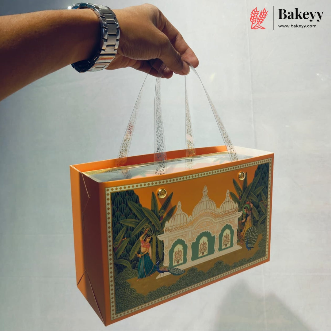 A hand holds a decorative orange bakery gift bag with clear plastic handles. The bag features a traditional Indian-style illustration of a white domed building, green foliage, and a peacock.