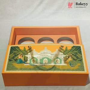 An orange rectangular gift box with a hinged lid, shown open on a light fabric background. The lid features a detailed traditional Indian illustration with a white domed palace, green foliage, peacocks, and a woman in a blue sari.