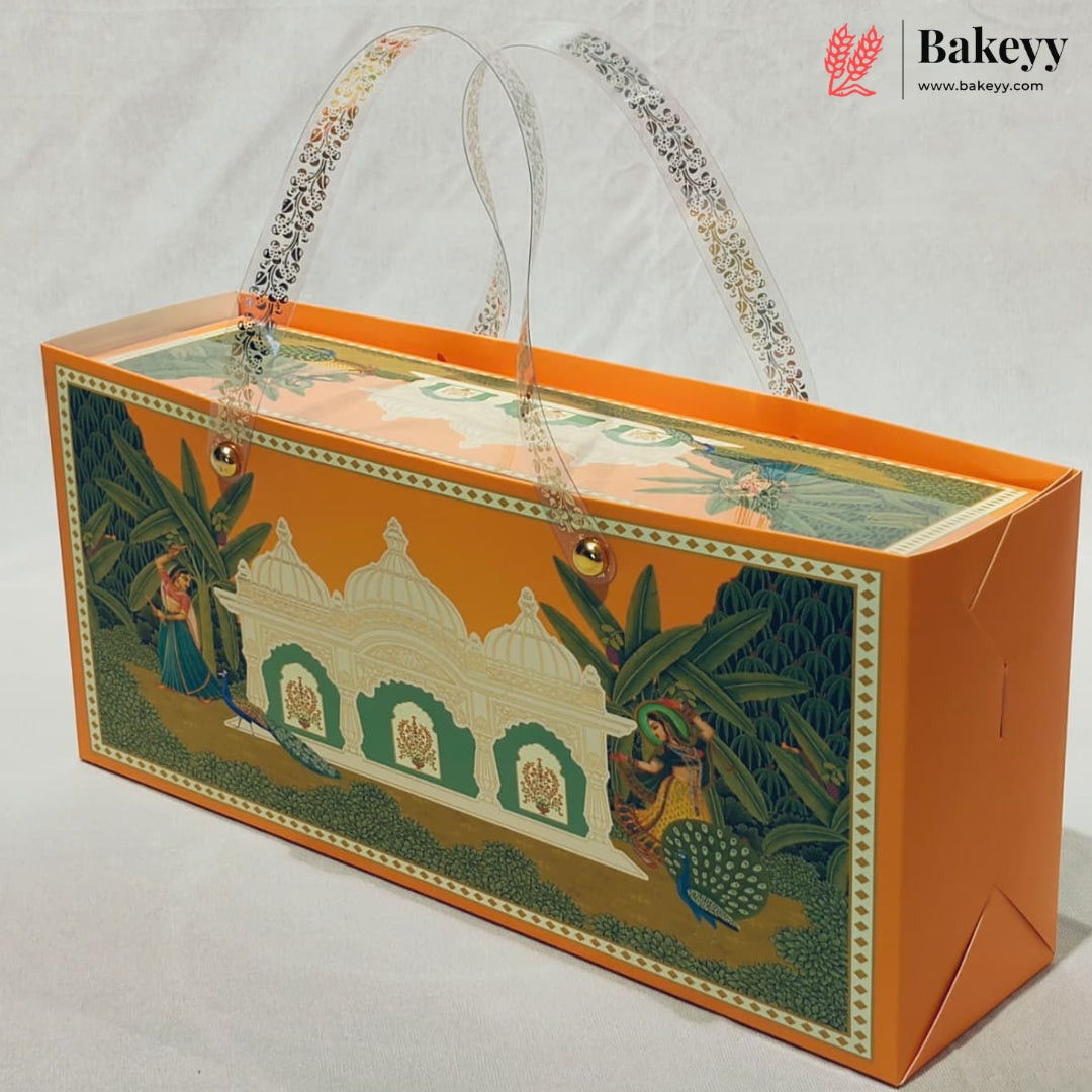 An orange bakery gift box with a traditional Indian illustration of a white domed building, green foliage, and peacocks on the front panel. The box features clear patterned paper handles and is photographed on a light gray background with a Bakeyy logo in the top right corner.