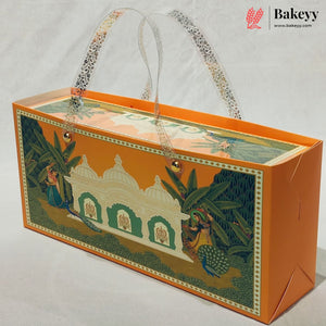 An orange bakery gift box with a traditional Indian illustration of a white domed building, green foliage, and peacocks on the front panel. The box features clear patterned paper handles and is photographed on a light gray background with a Bakeyy logo in the top right corner.