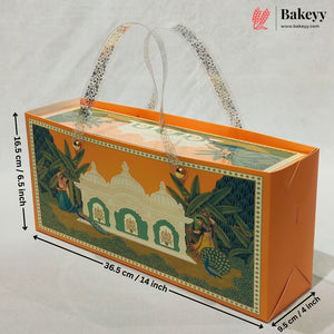 An orange rectangular bakery gift box with clear plastic handles and a colorful illustrated front panel showing a domed building, green foliage, and peacocks. Black dimension arrows and text show the box measurements: 36.